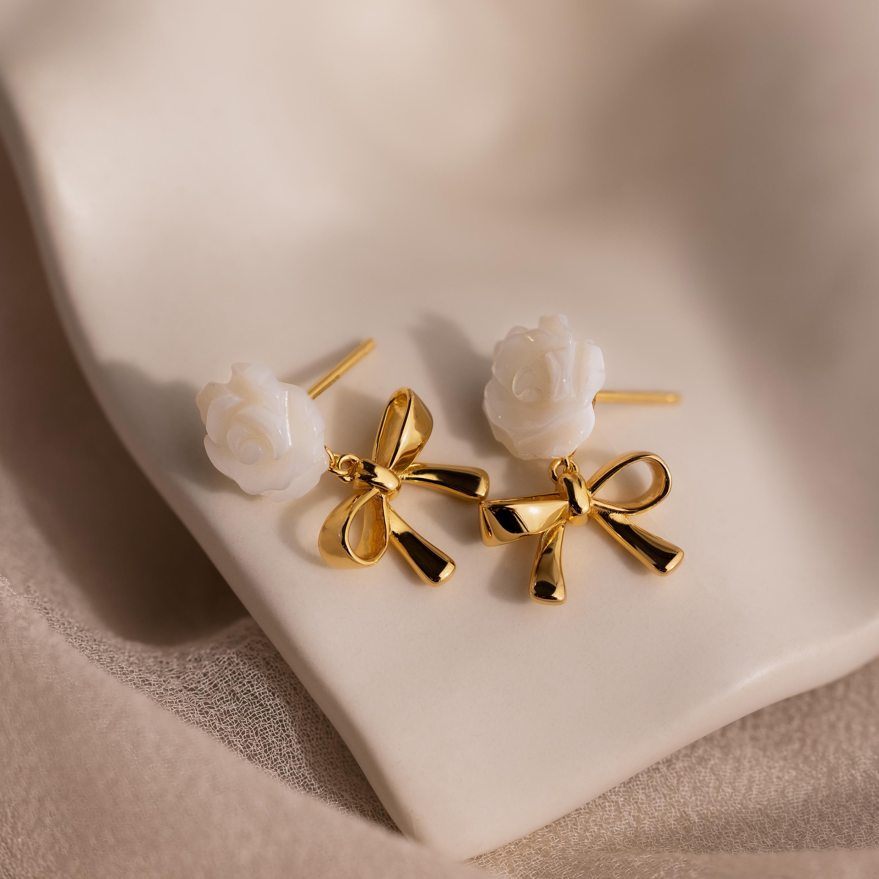 Close-up of white rose and gold bow earrings with posts visible, resting on ivory ceramic.