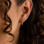 Close-up of a woman's ear with curly hair, wearing an elegant pair of Lace Diamond Huggies—small, dangling gold earrings adorned with a sparkling gemstone.