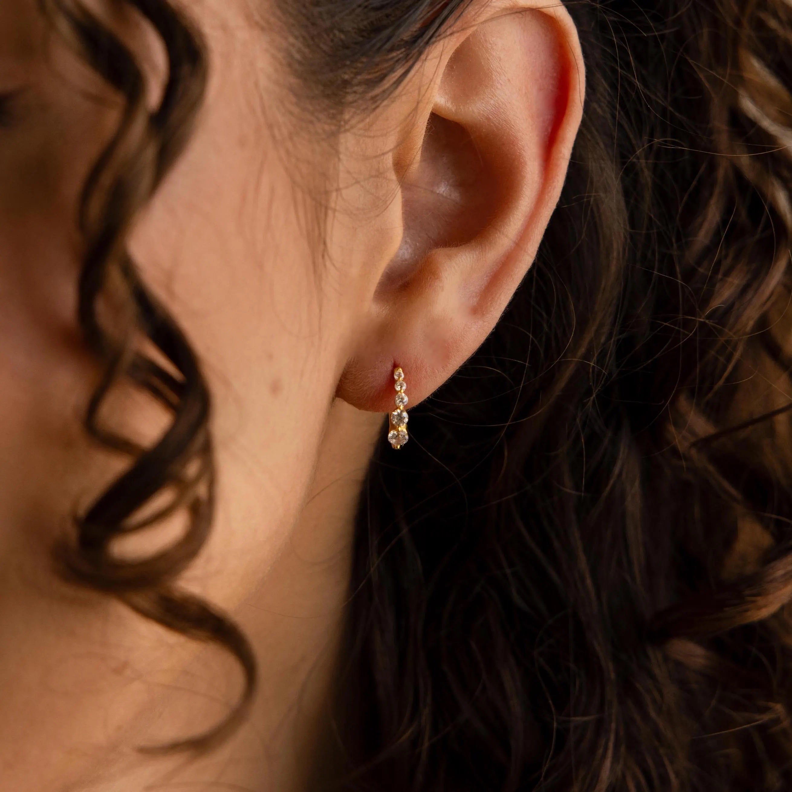 Close-up of a woman's ear with curly hair, wearing an elegant pair of Lace Diamond Huggies—small, dangling gold earrings adorned with a sparkling gemstone.