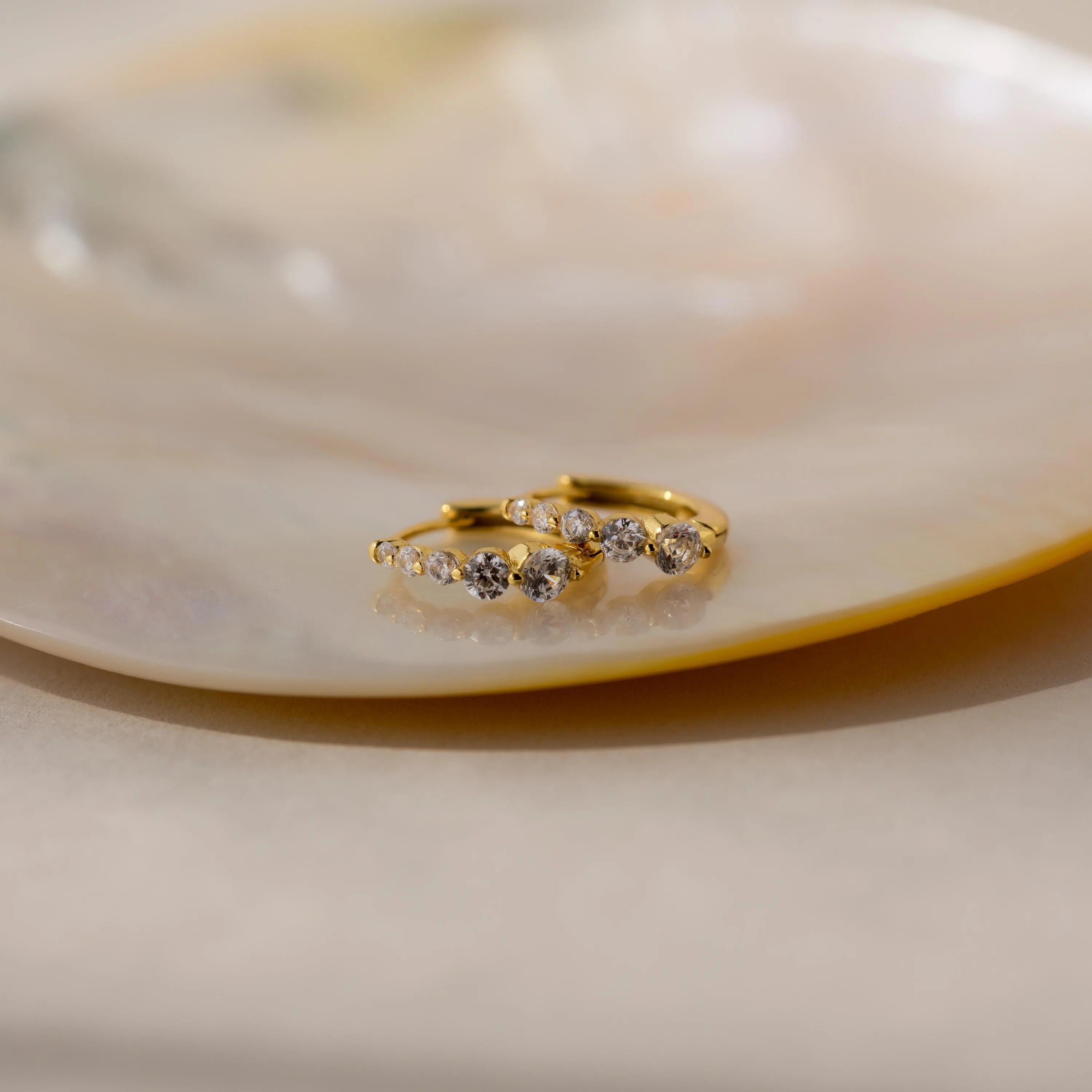 Lace Diamond Huggies—gold hoop earrings adorned with small clear gemstones—are elegantly displayed on a glossy, light-colored dish.