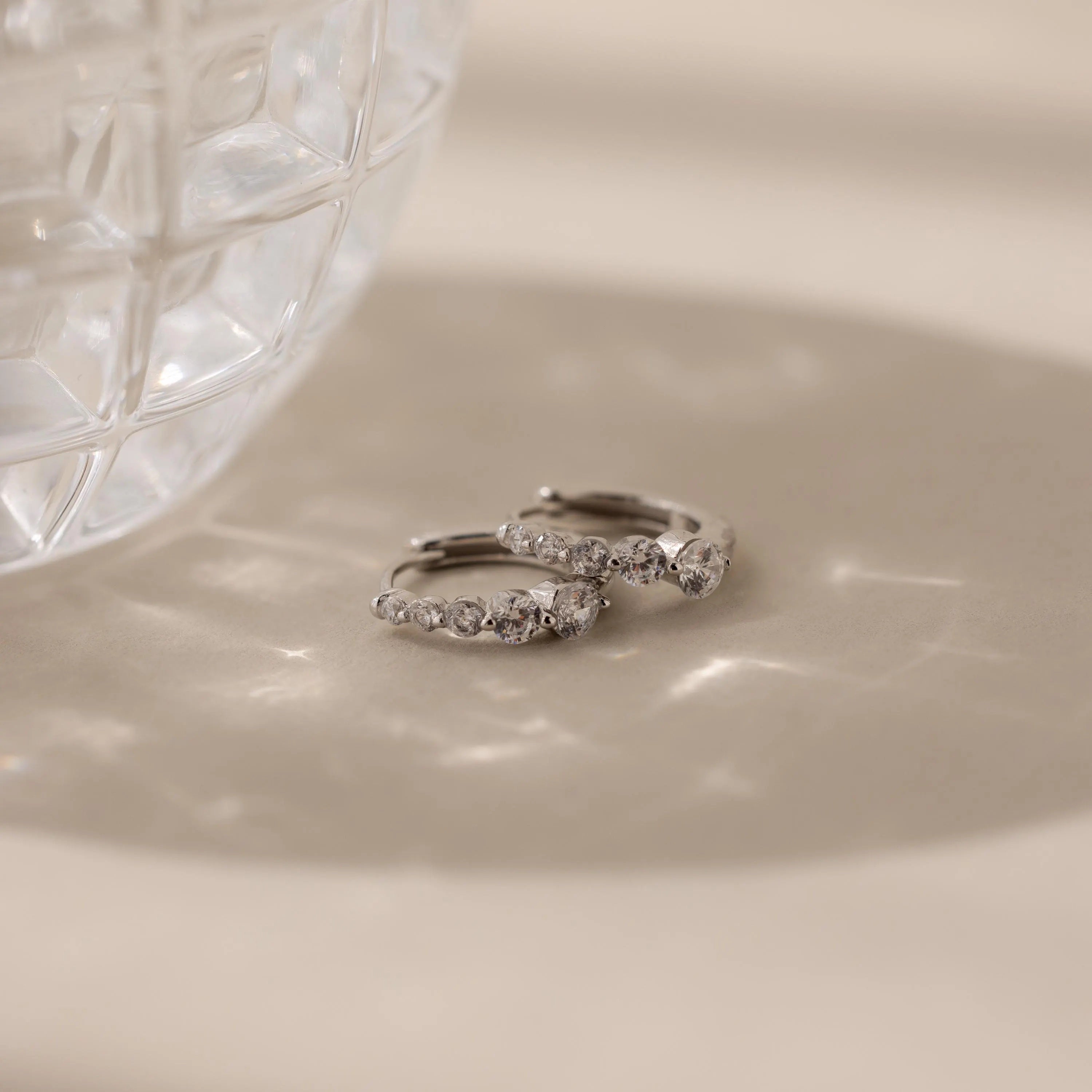 Two elegant earrings—Lace Diamond Huggies—sit beside a faceted glass, casting delicate shadows on a beige surface.