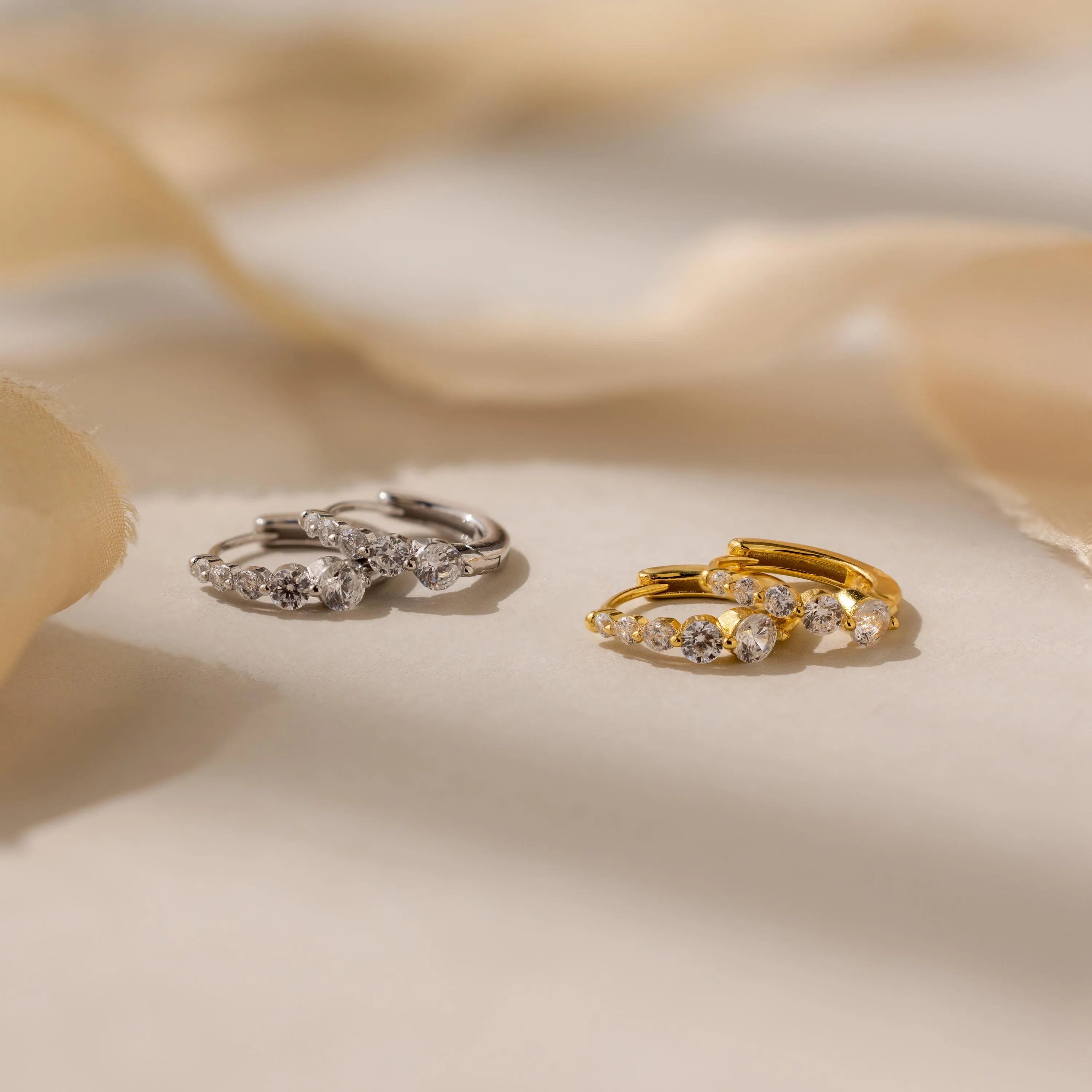 Two pairs of Lace Diamond Huggies—one silver, one gold—each adorned with three sparkling stones, radiate timeless elegance on a soft beige surface.