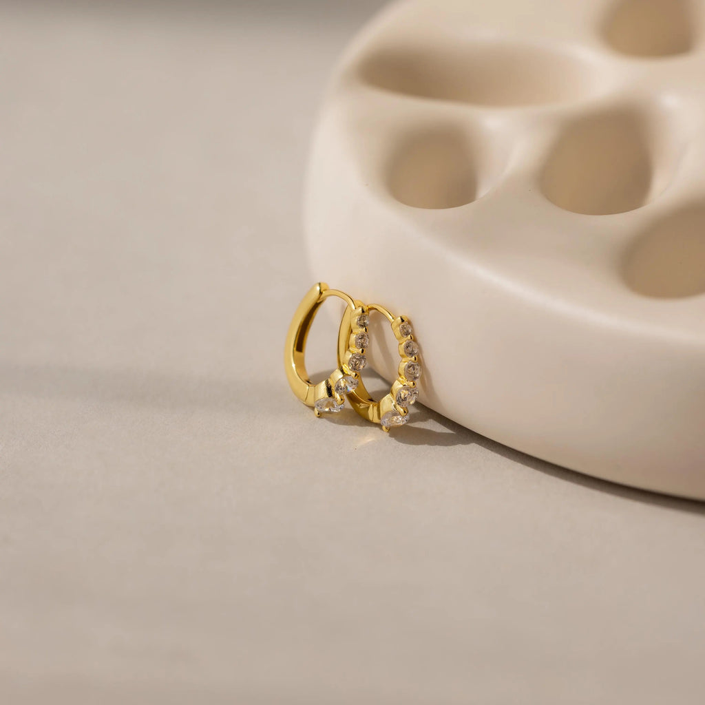 The Lace Diamond Huggies, elegant earrings with embedded crystals, rest against a beige curved ceramic dish, echoing the timeless style of classic diamond hoops.