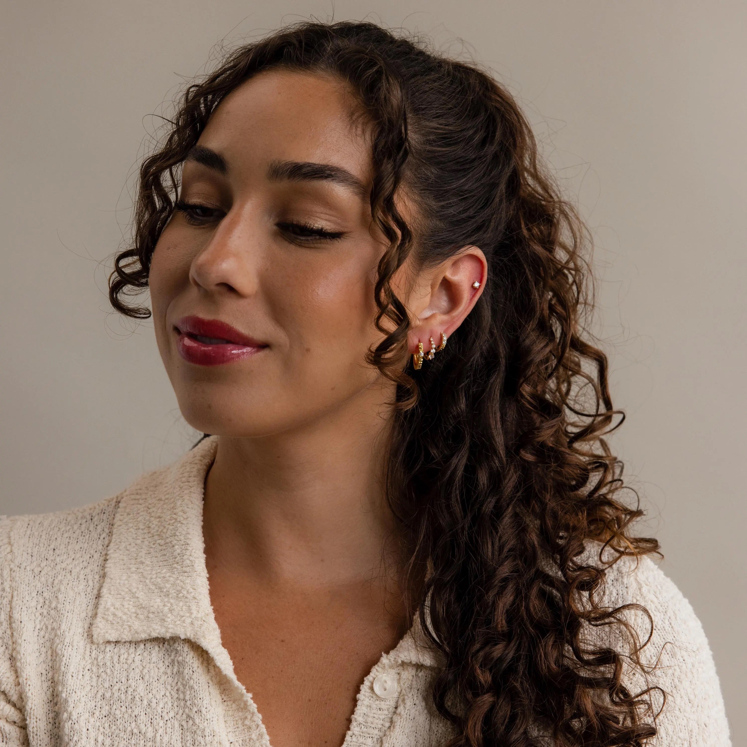 Woman with curly brown hair in a ponytail, wearing elegant Lace Diamond Huggies and a cream textured top, looking down.