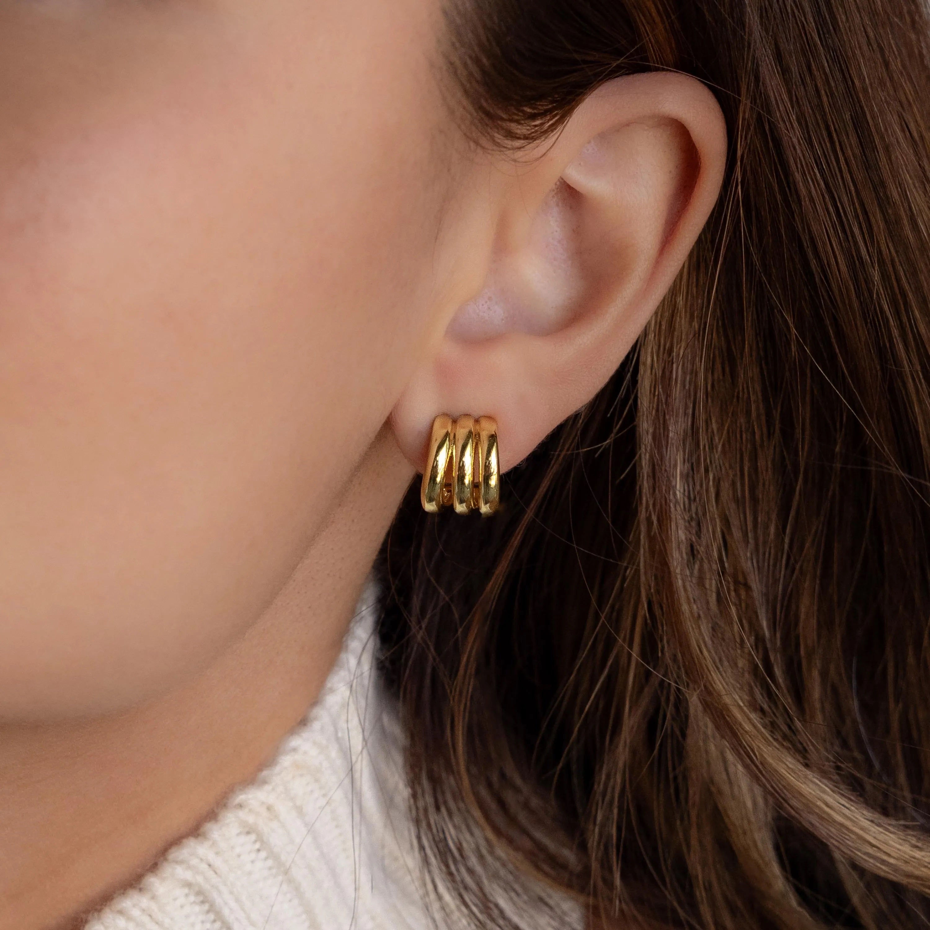 Close-up of a woman's ear wearing the Triple Loop Huggie Earrings in gold, styled with brown hair and a white knit sweater—an ideal accessory for everyday wear.