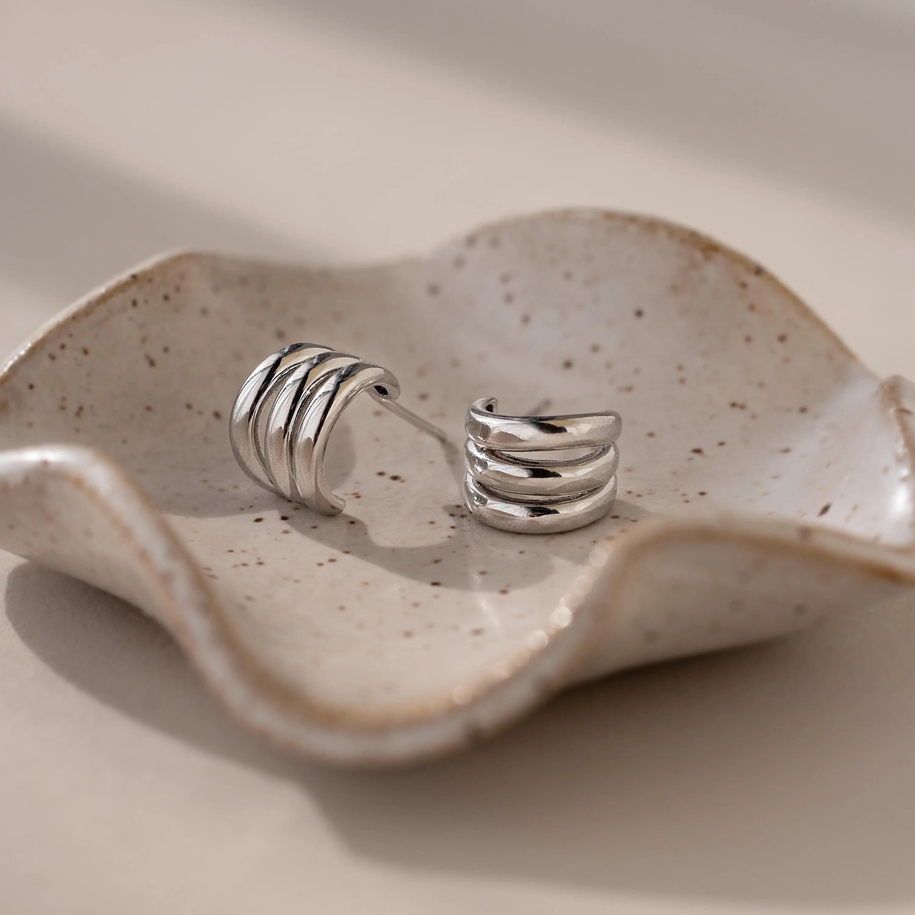 Triple Loop Huggie Earrings rest on a wavy, speckled ceramic dish in soft natural light—ideal for stacked ear styling or effortless everyday wear.