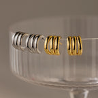 Two pairs of Triple Loop Huggie Earrings—one silver, one gold—displayed on a glass dish rim. Ideal for stacked ear looks and easy everyday style.