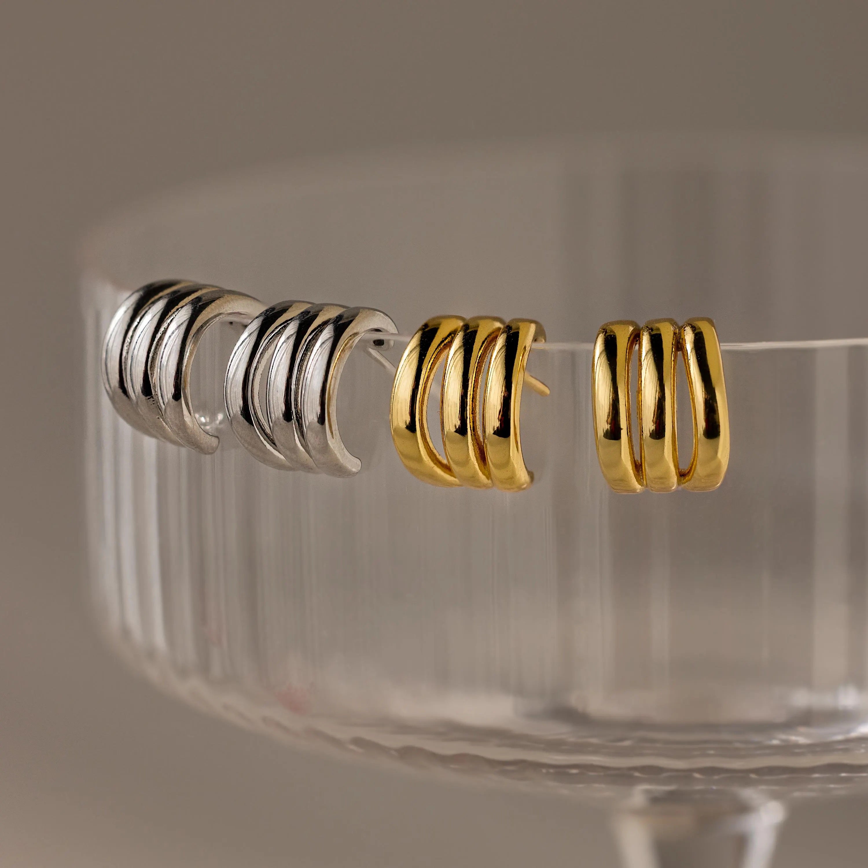 Two pairs of Triple Loop Huggie Earrings—one silver, one gold—displayed on a glass dish rim. Ideal for stacked ear looks and easy everyday style.