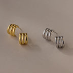 Two pairs of Triple Loop Huggie Earrings, one gold and one silver, ideal for stacking or daily wear, shown on a beige surface.