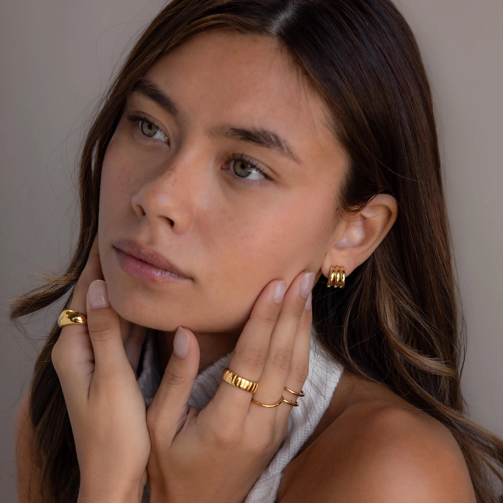 A woman with long brown hair wears Triple Loop Huggie Earrings in a stacked style, paired with gold rings, as she touches her face and gazes thoughtfully—showcasing these earrings as the perfect accessory for everyday wear.