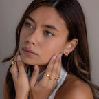 A woman with long brown hair wears Triple Loop Huggie Earrings in a stacked style, paired with gold rings, as she touches her face and gazes thoughtfully—showcasing these earrings as the perfect accessory for everyday wear.