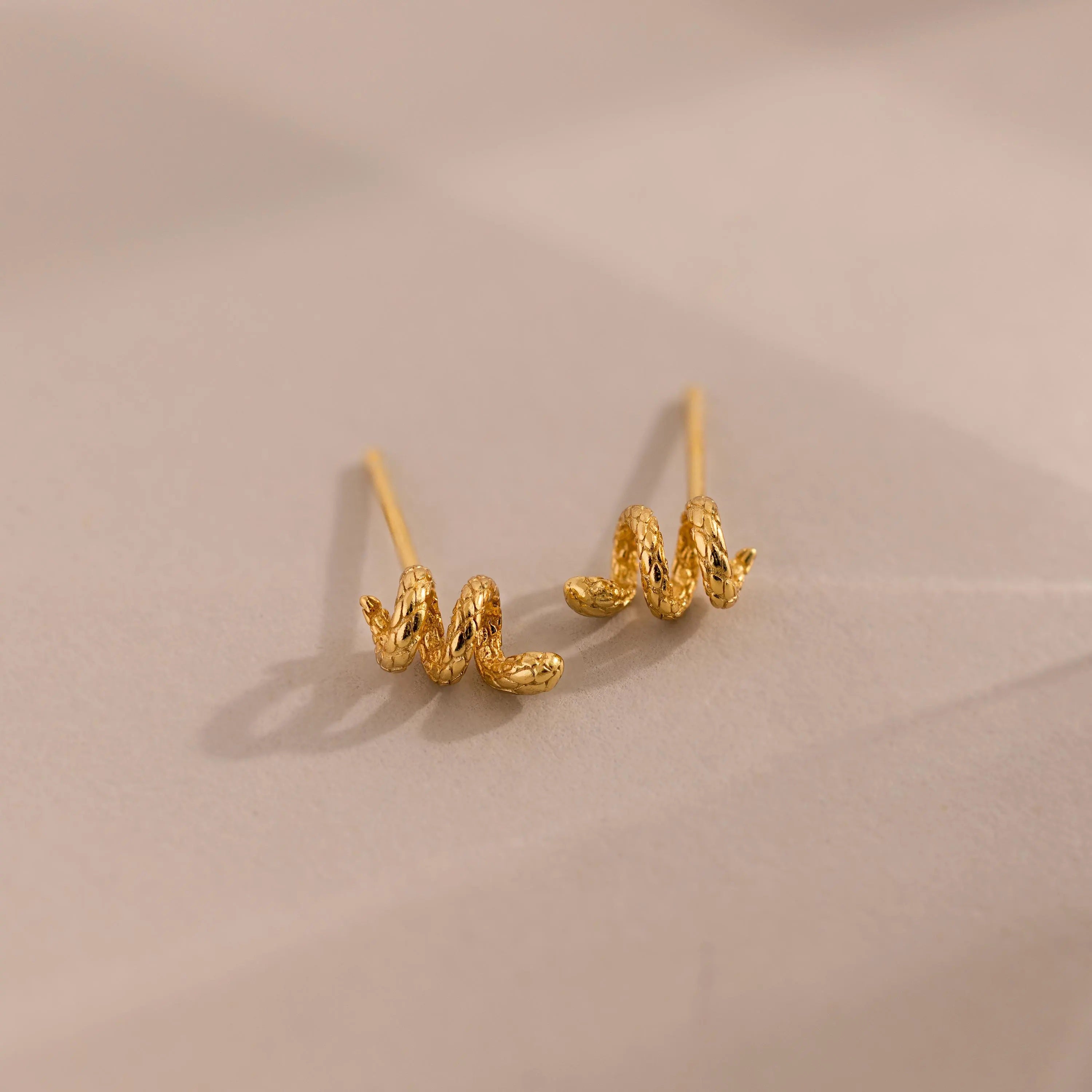 Gold 3D twisted snake stud earrings resting against a neutral surface.
