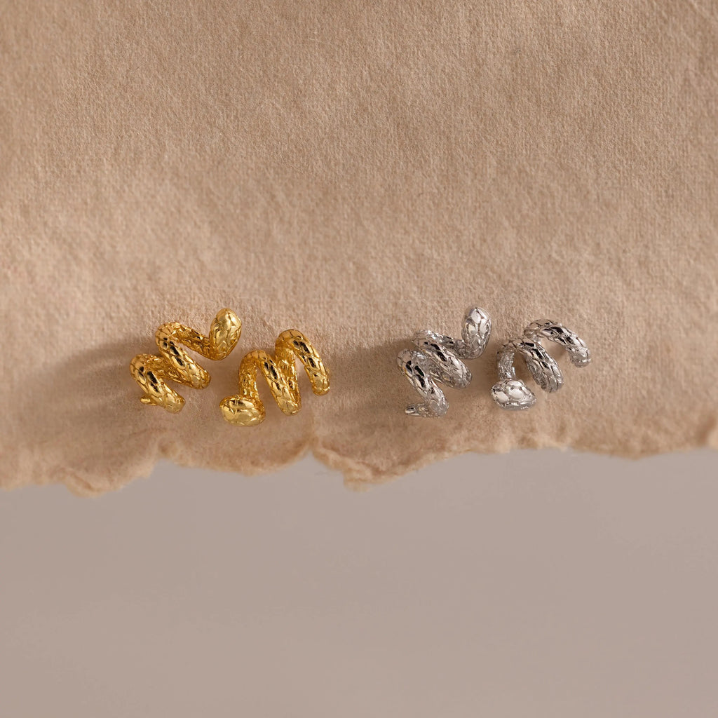 Product display of two pairs of Twisted Snake Earrings – one in gold and one in silver – laid out side by side on a rippled beige fabric background. Both versions showcase the same serpentine coil design with textured scale details