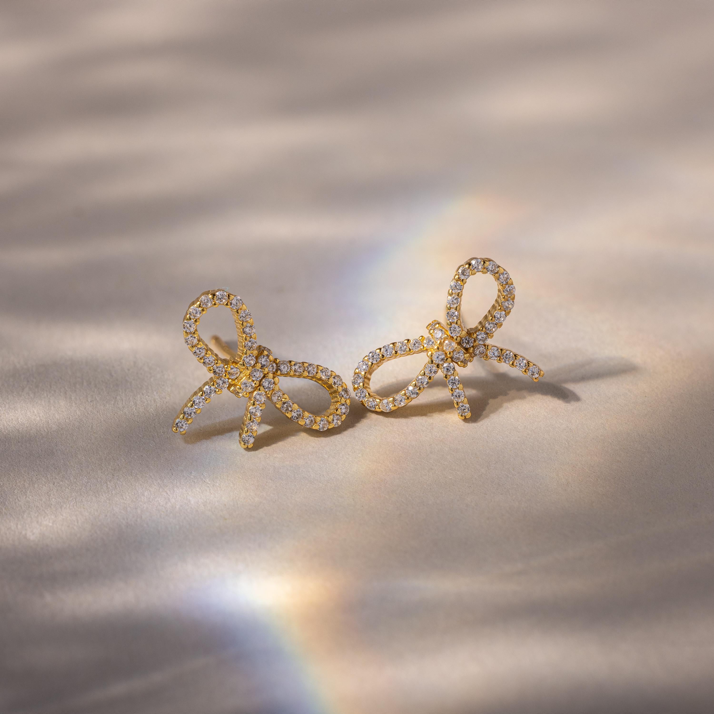 Gold pavé bow stud earrings with sparkling diamond stones, resting on a neutral surface with a subtle rainbow light reflection.