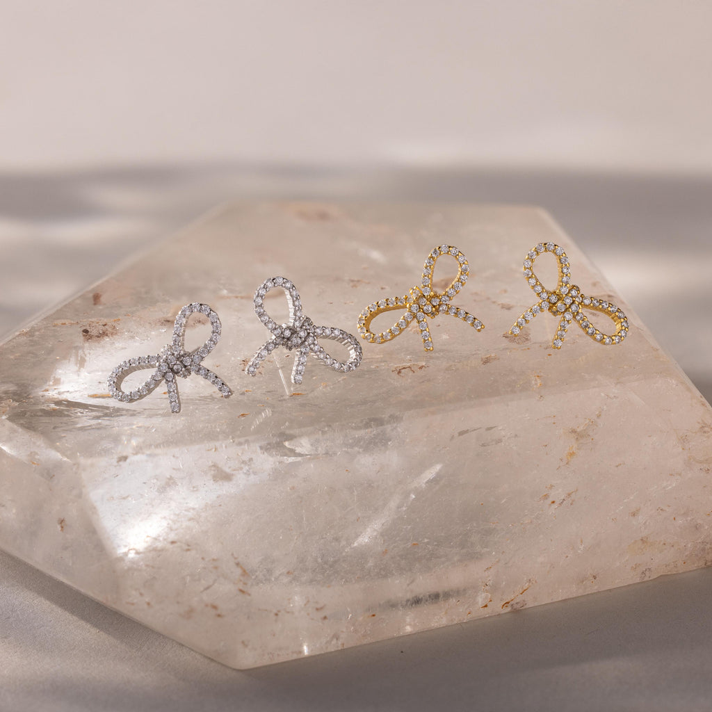 Set of gold and silver pavé bow stud earrings displayed on a clear quartz crystal slab, showcasing their sparkling ribbon design under soft natural light.