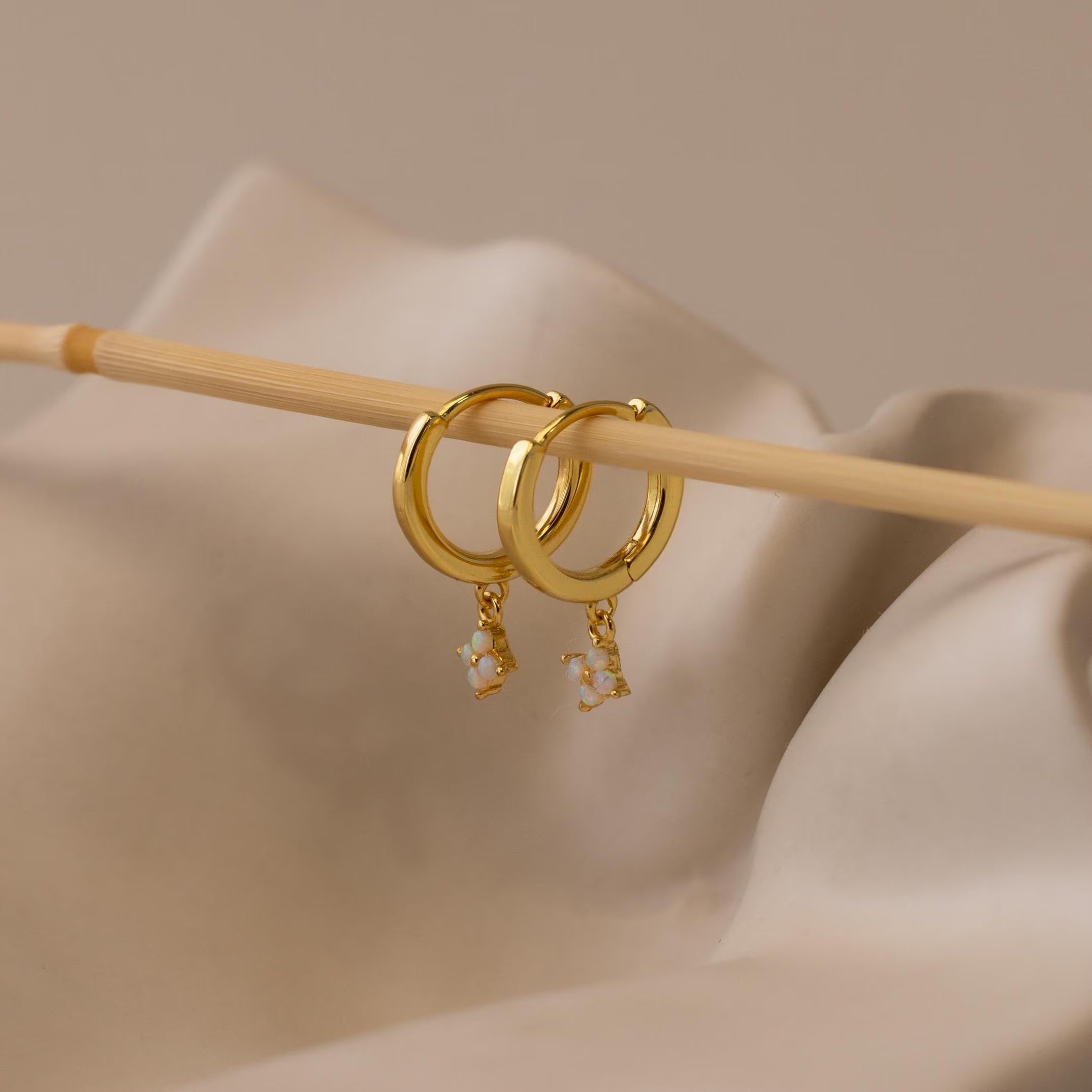 The Ivy Birthstone Hoops, featuring two gold hoops with small birthstone charms, hang on a thin wooden stick against a neutral background, offering a perfect option for personalized jewelry.