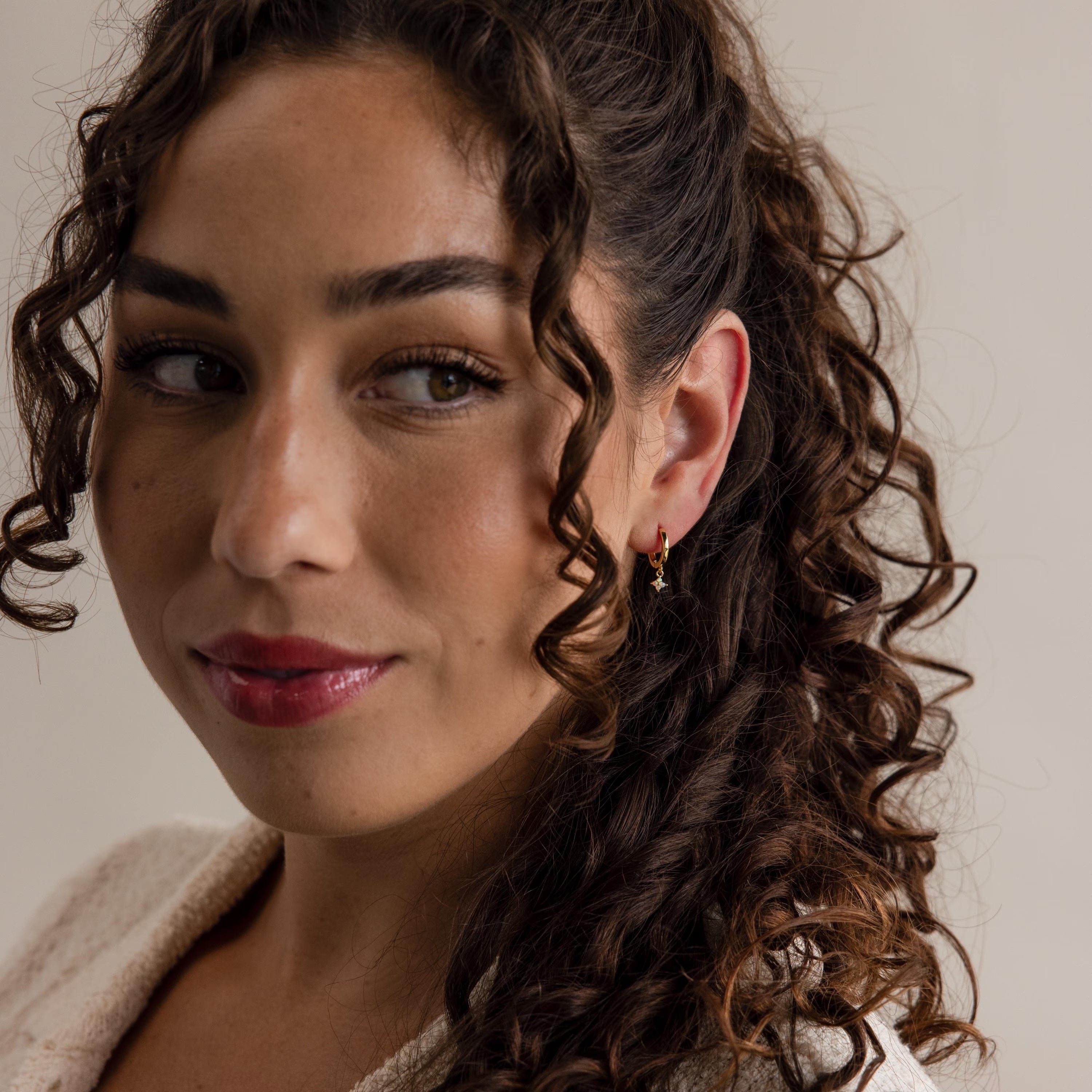 Woman with long curly hair, looking slightly left, wearing Ivy Birthstone Hoops and light-colored clothing.