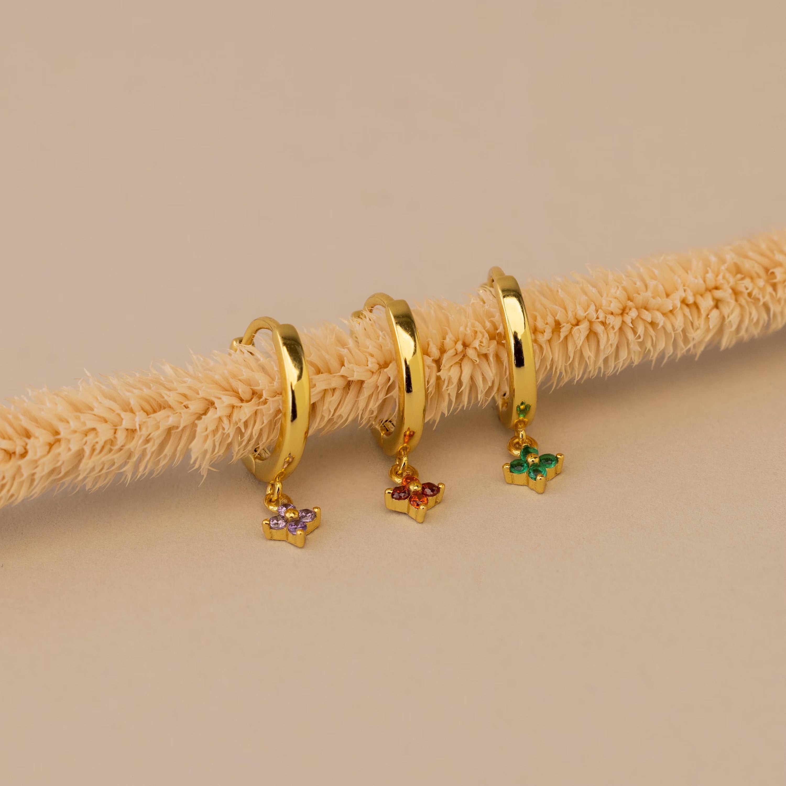 Three Ivy Birthstone Hoops, each featuring gold hoops with vibrant butterfly charms, are displayed on a beige textured rod against a neutral background—perfect as personalized jewelry.