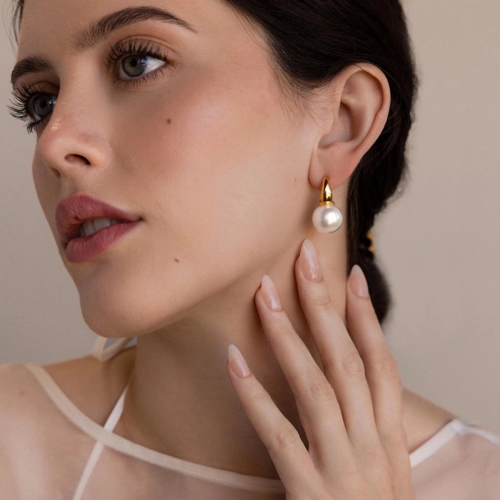 A fair-skinned woman wears Dome Pearl Huggies, elegant gold and pearl hoop earrings, gently touching her neck with manicured fingers.
