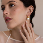 A fair-skinned woman wears Dome Pearl Huggies, elegant gold and pearl hoop earrings, gently touching her neck with manicured fingers.