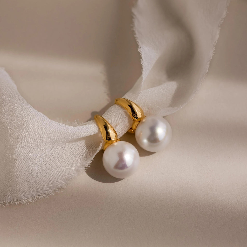 Dome Pearl Huggies shine with gold and pearl accents, displayed on soft white fabric and illuminated by gentle natural light and shadows.
