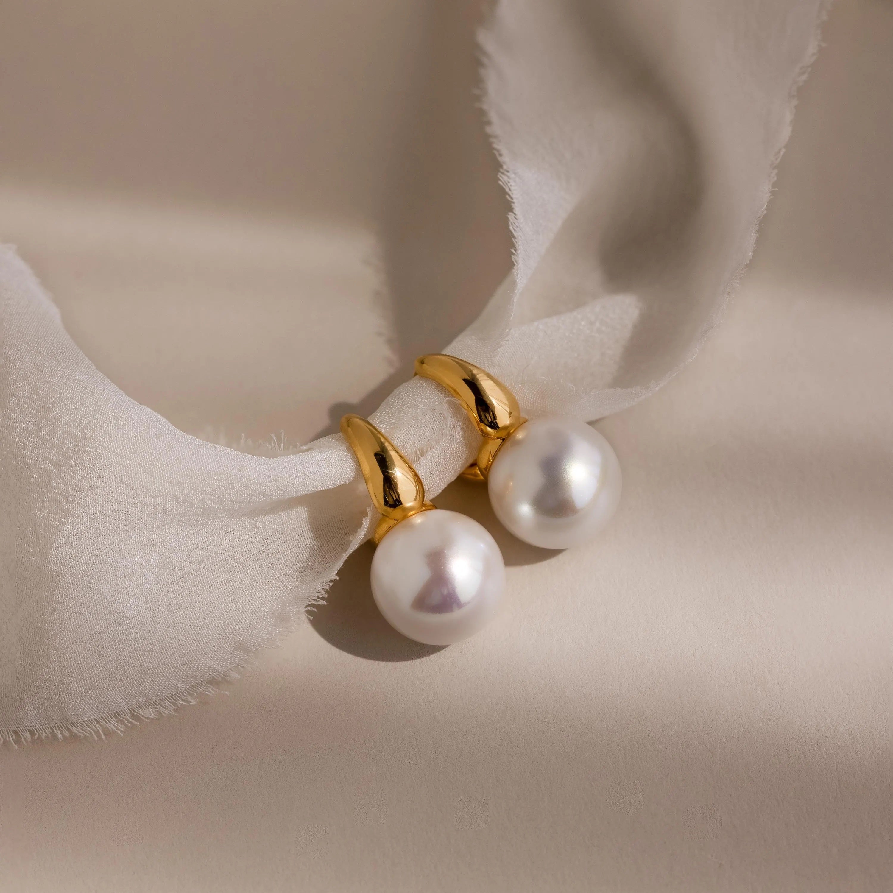 Dome Pearl Huggies shine with gold and pearl accents, displayed on soft white fabric and illuminated by gentle natural light and shadows.