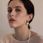 A woman with dark hair wearing Dome Pearl Huggies—elegant hoop earrings featuring a pearl gemstone—looks at the camera with a neutral expression.