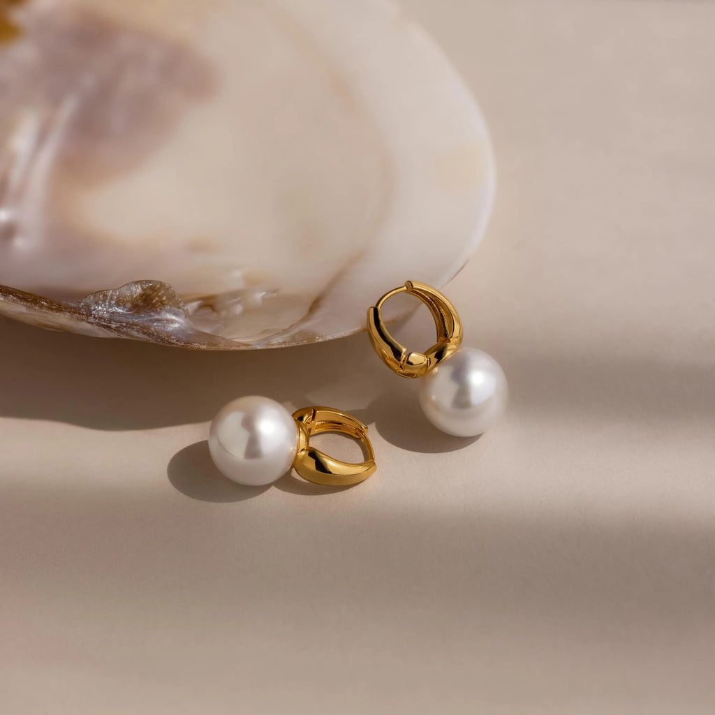 The Dome Pearl Huggies rest beside an open seashell on a soft beige background.