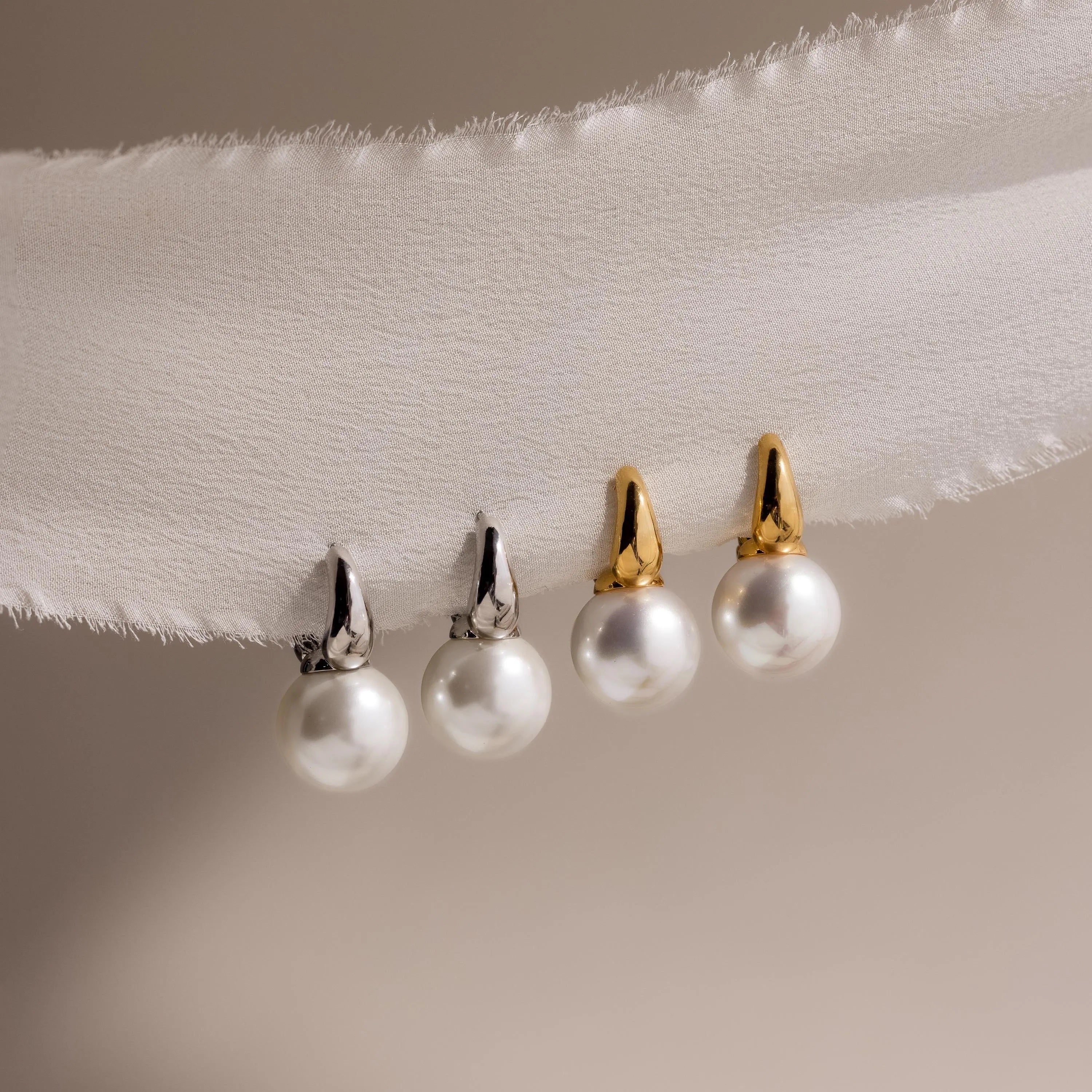 Two pairs of pearl gemstone earrings, featuring Dome Pearl Huggies with silver hooks and classic hoop earrings with gold hooks, are displayed on a white fabric strip.