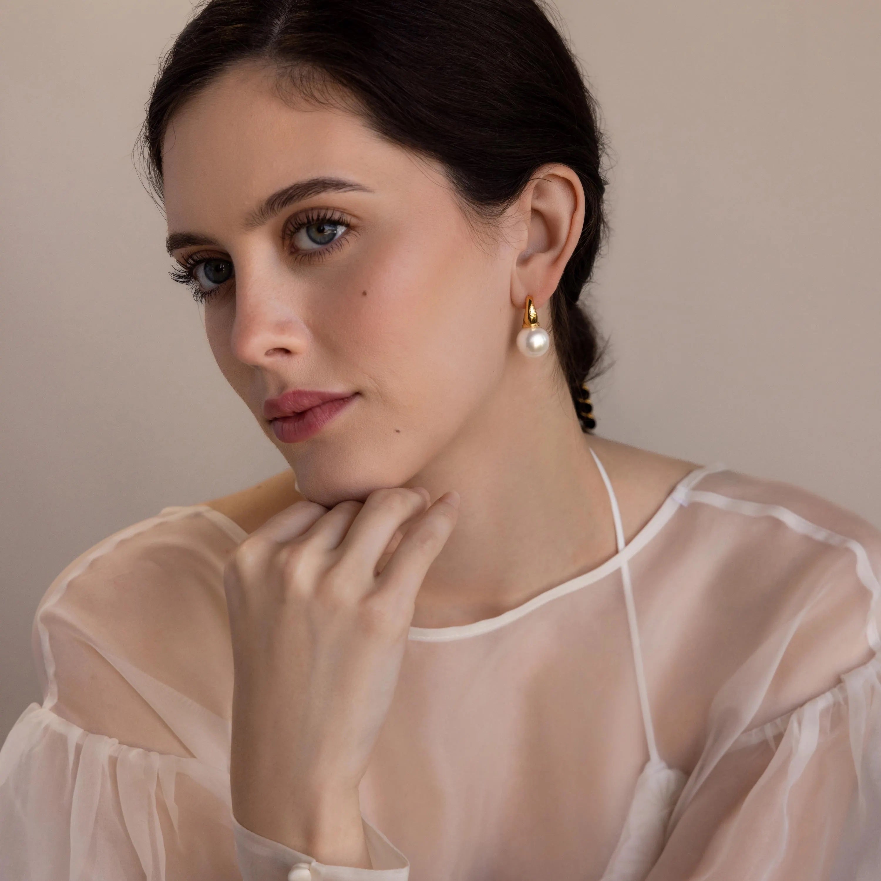 A woman with dark hair, dressed in a sheer white top, rests her chin on her hand while wearing elegant Dome Pearl Huggies.