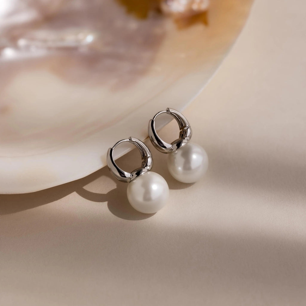 Dome Pearl Huggies, silver hoop earrings accented with round pearl gemstones, rest on a soft beige surface beside a shell.