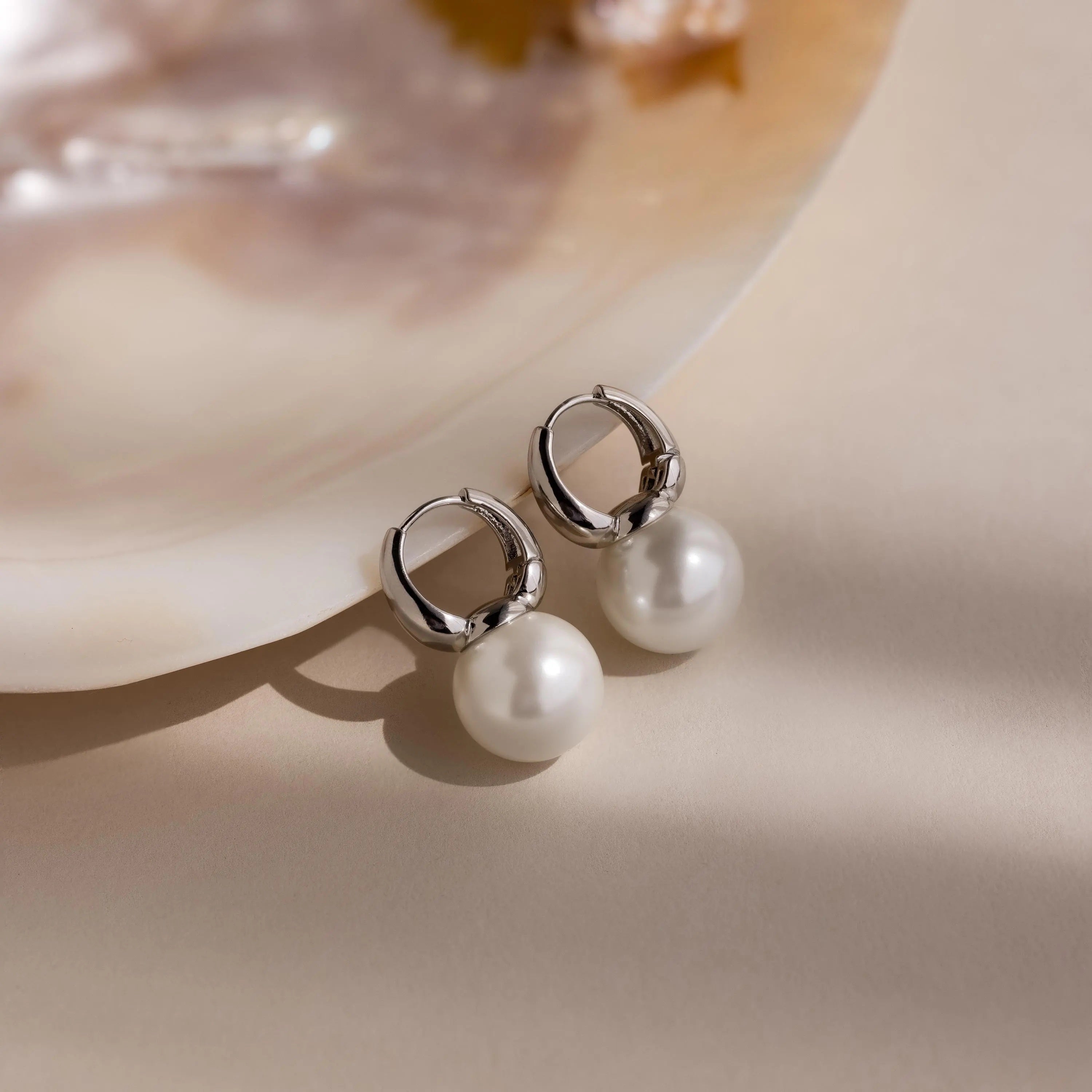 Dome Pearl Huggies, silver hoop earrings accented with round pearl gemstones, rest on a soft beige surface beside a shell.