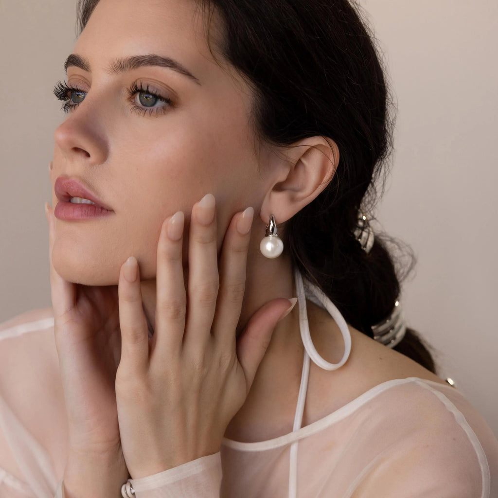 A woman with dark hair wears Dome Pearl Huggies and a sheer white top, gently touching her face in a thoughtful pose.