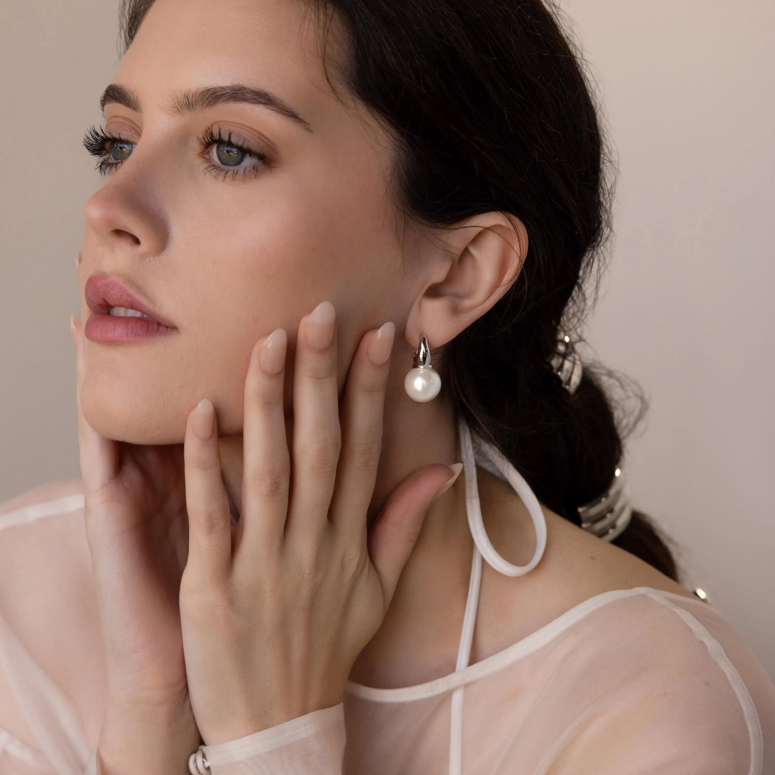 A woman with dark hair wears Dome Pearl Huggies and a sheer white top, gently touching her face in a thoughtful pose.