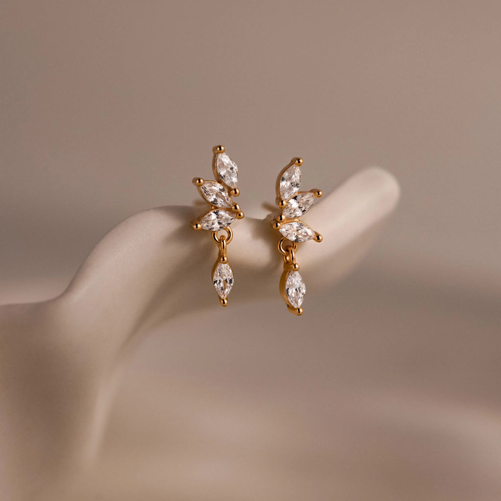 Close-up of dainty gold CZ diamond drop earrings with four marquise-cut stones, displayed on a neutral surface.