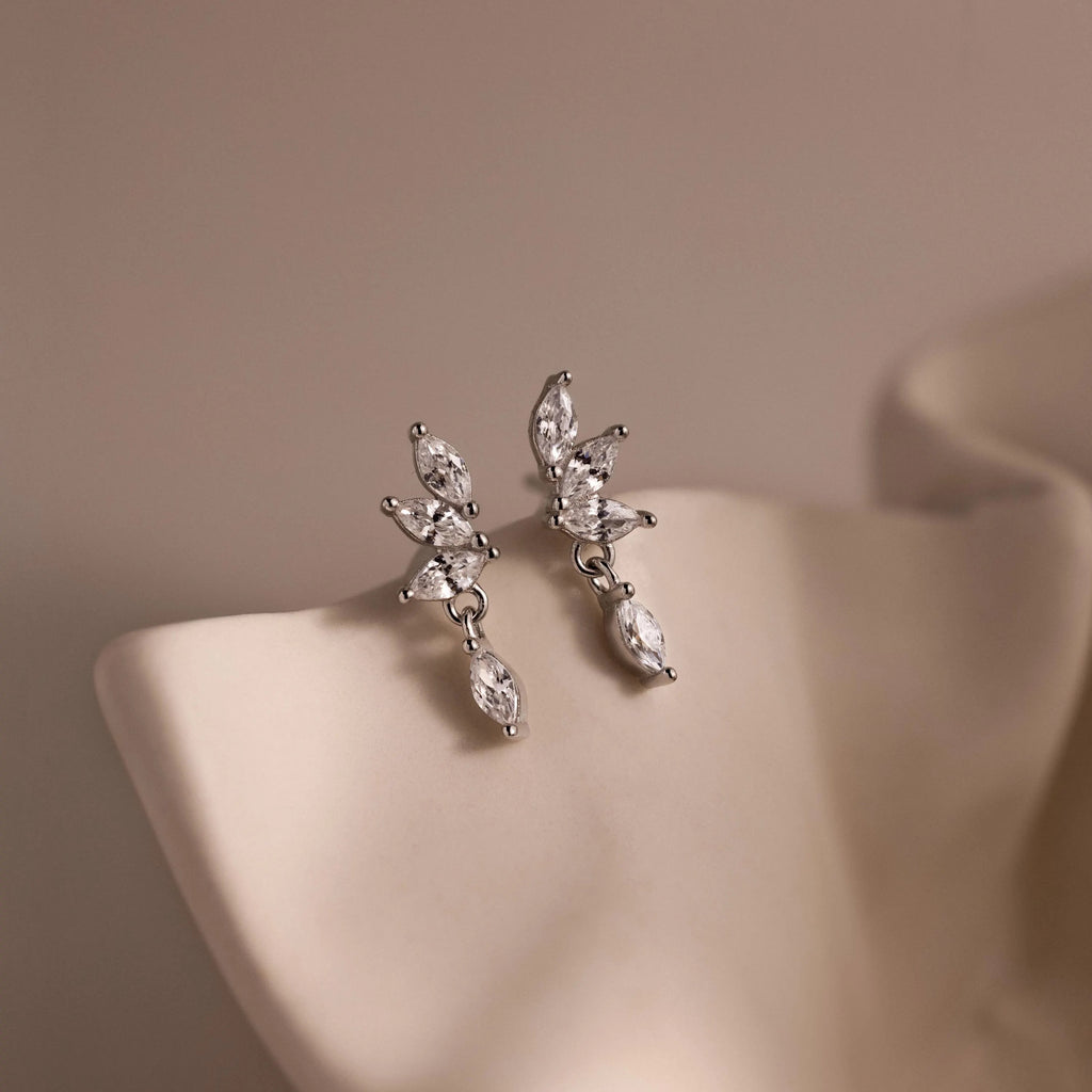 Close-up of dainty silver CZ diamond drop earrings with four marquise-cut stones, displayed on a neutral surface.