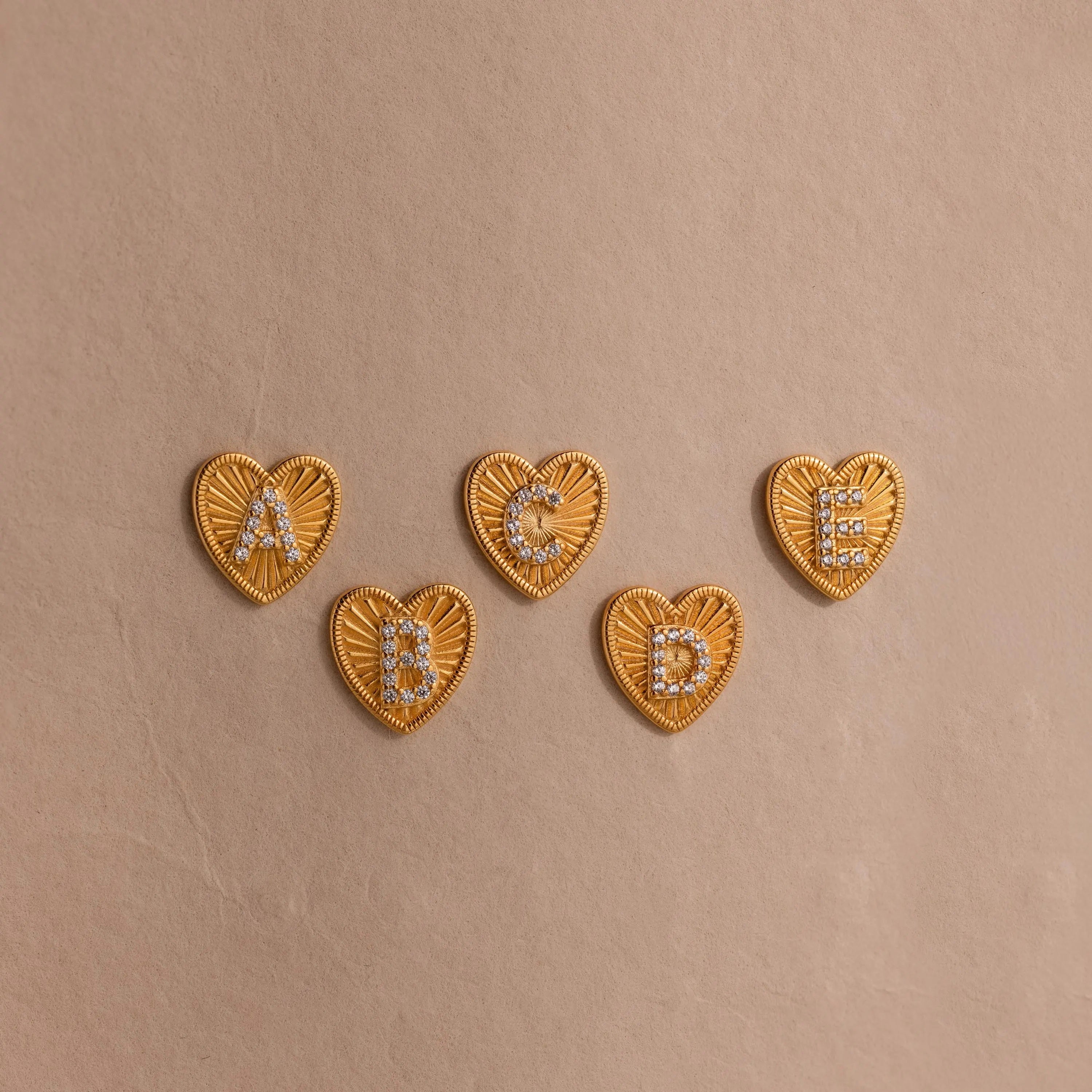 5 gold Ribbed Heart Initial Stud Earrings with the letters A, B, C, D, and E in pave detailing, displayed on a neutral background.