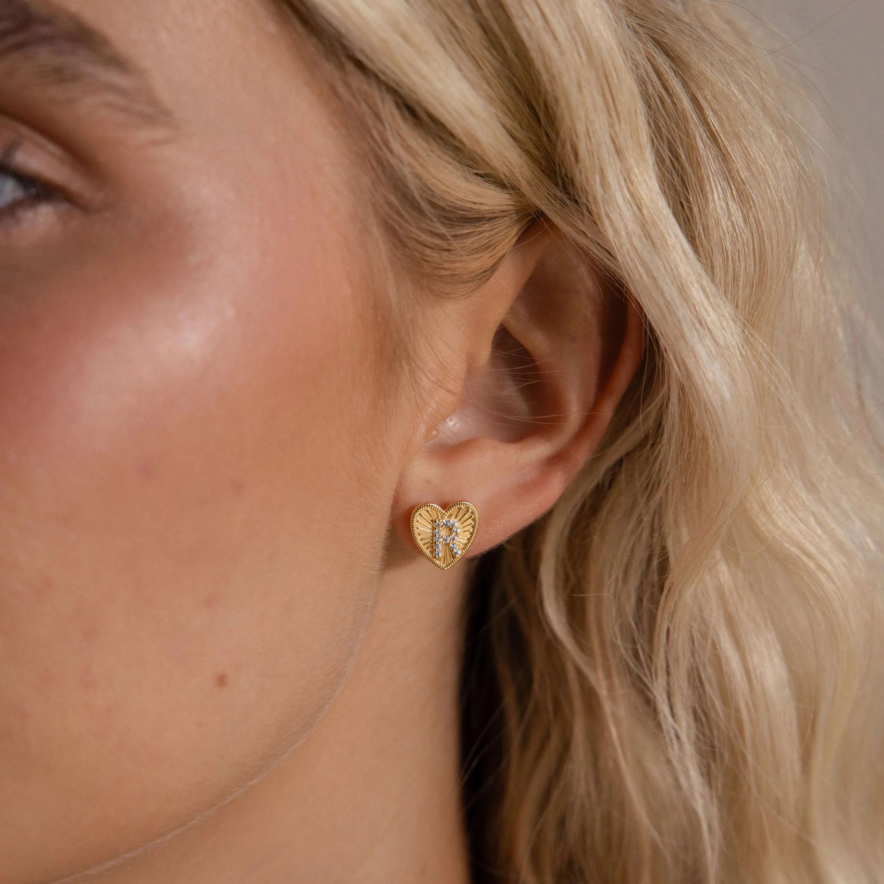 Model wearing a gold Ribbed Heart Initial Stud Earring with the letter R in CZ pave detailing.