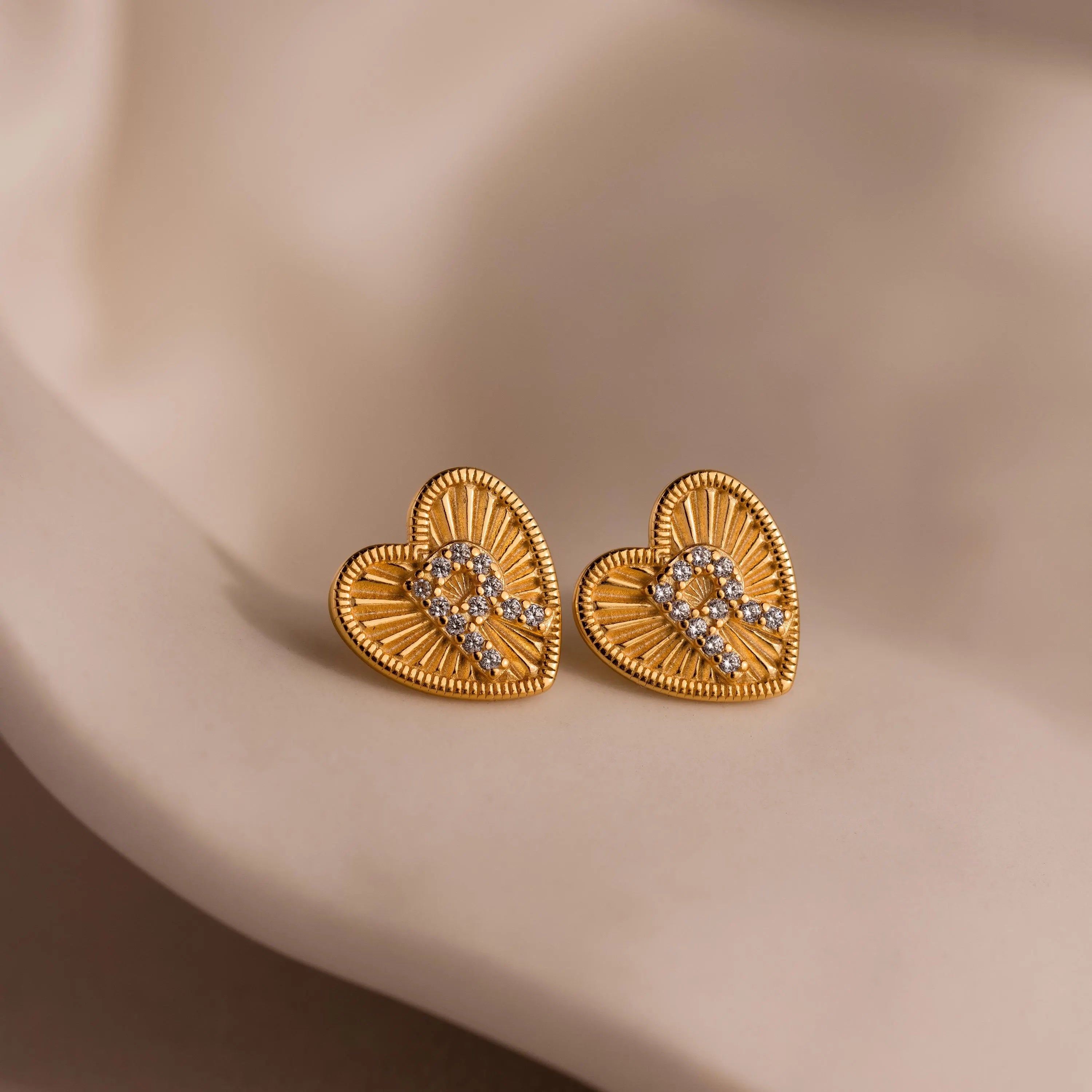Close-up of a pair of gold Ribbed Heart Initial Studs with the letter R in CZ pave detailing, resting against a neutral background.