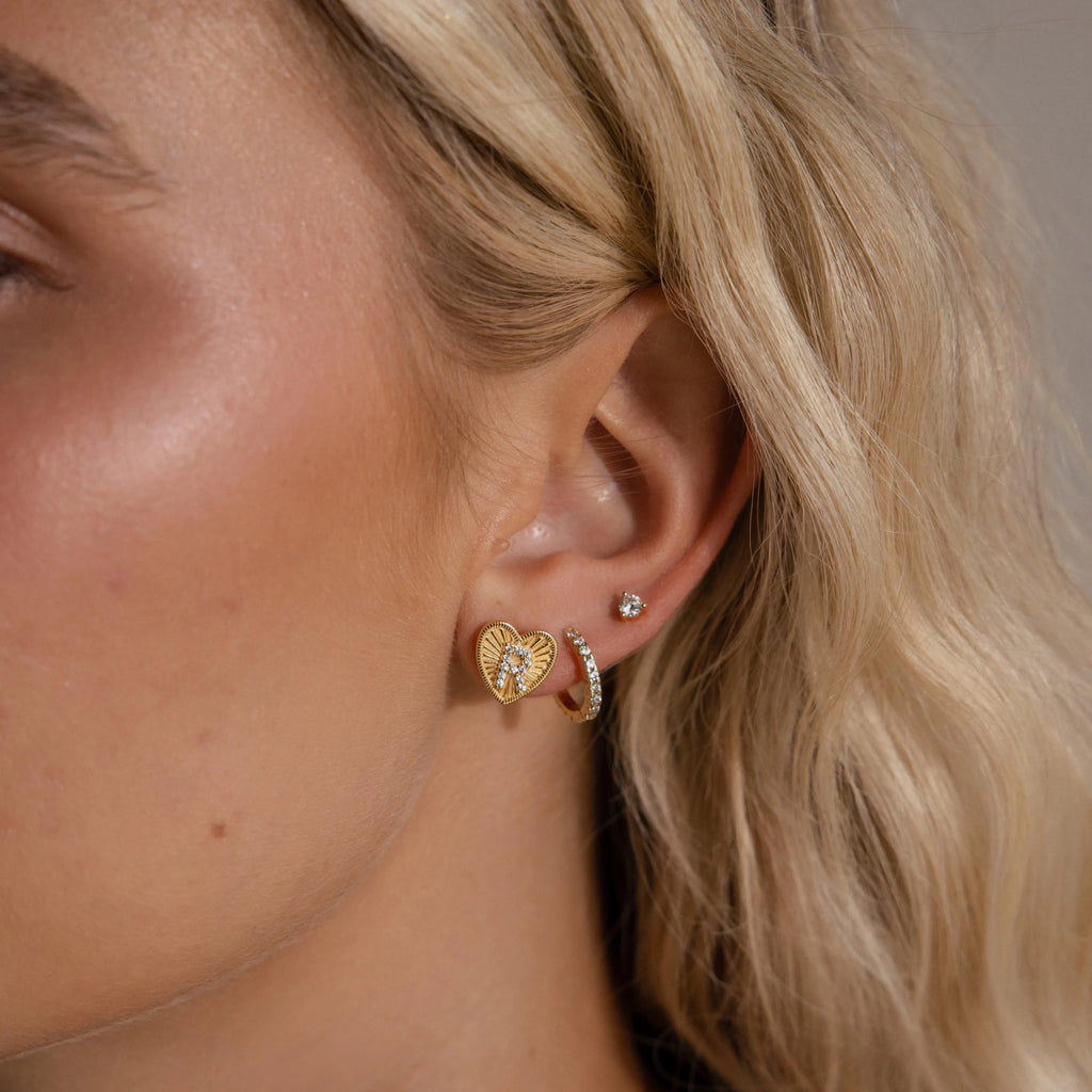 Model wearing a gold Ribbed Heart Initial Earring with a pave letter R, stacked with a pave huggie hoop and dainty diamond stud.