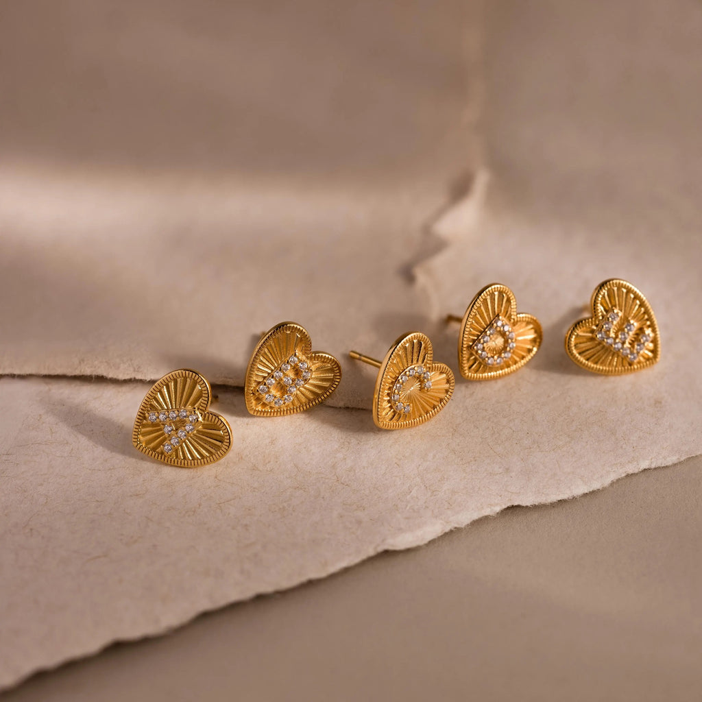 5 gold Ribbed Heart Initial Stud Earrings with sparkling A, B, C, D, and E pave letters displayed on textured neutral paper backdrop.