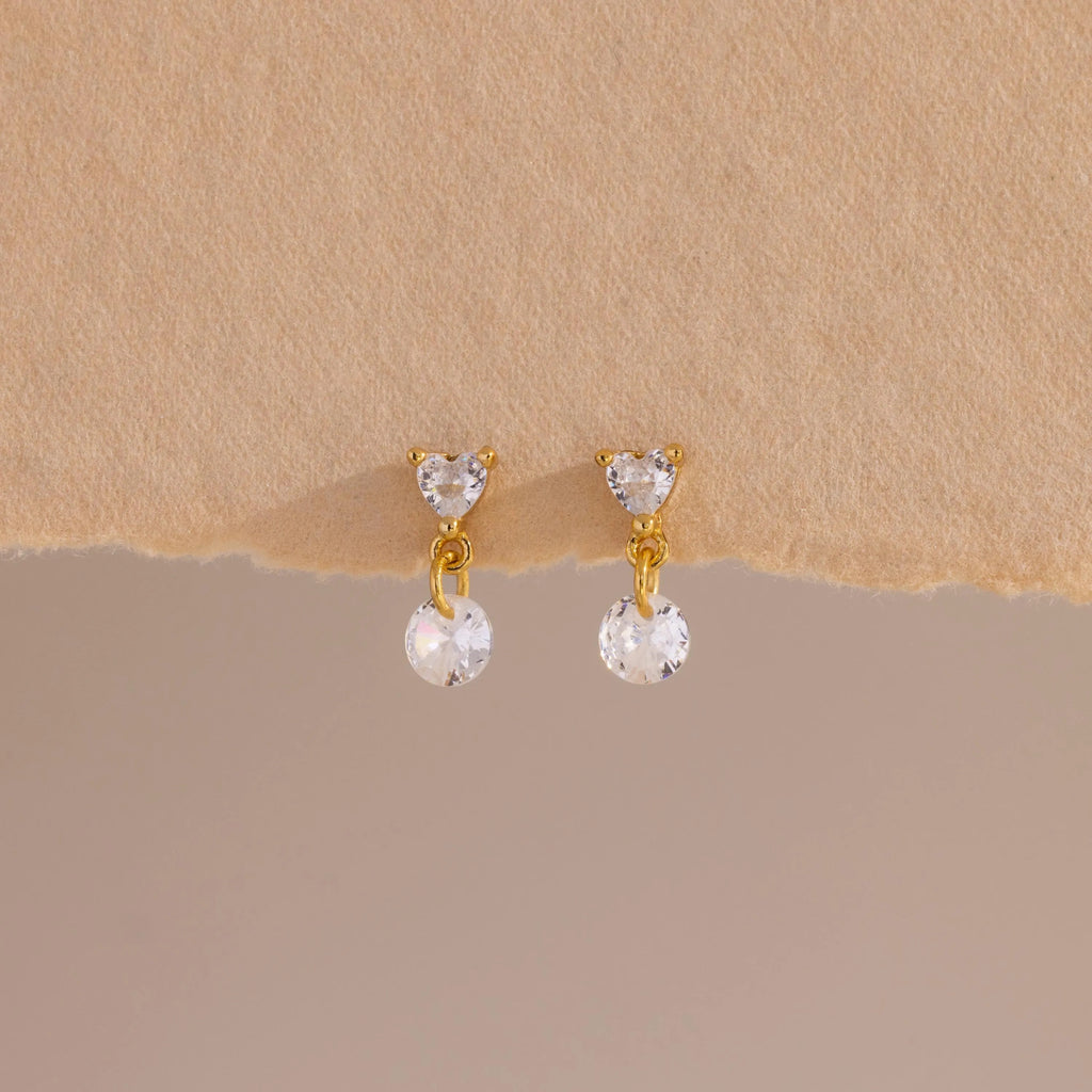 A close-up of a pair of 18k gold flat back earrings featuring heart-shaped studs with a dangling round crystal, displayed against a soft beige textured surface.