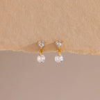 A close-up of a pair of 18k gold flat back earrings featuring heart-shaped studs with a dangling round crystal, displayed against a soft beige textured surface.