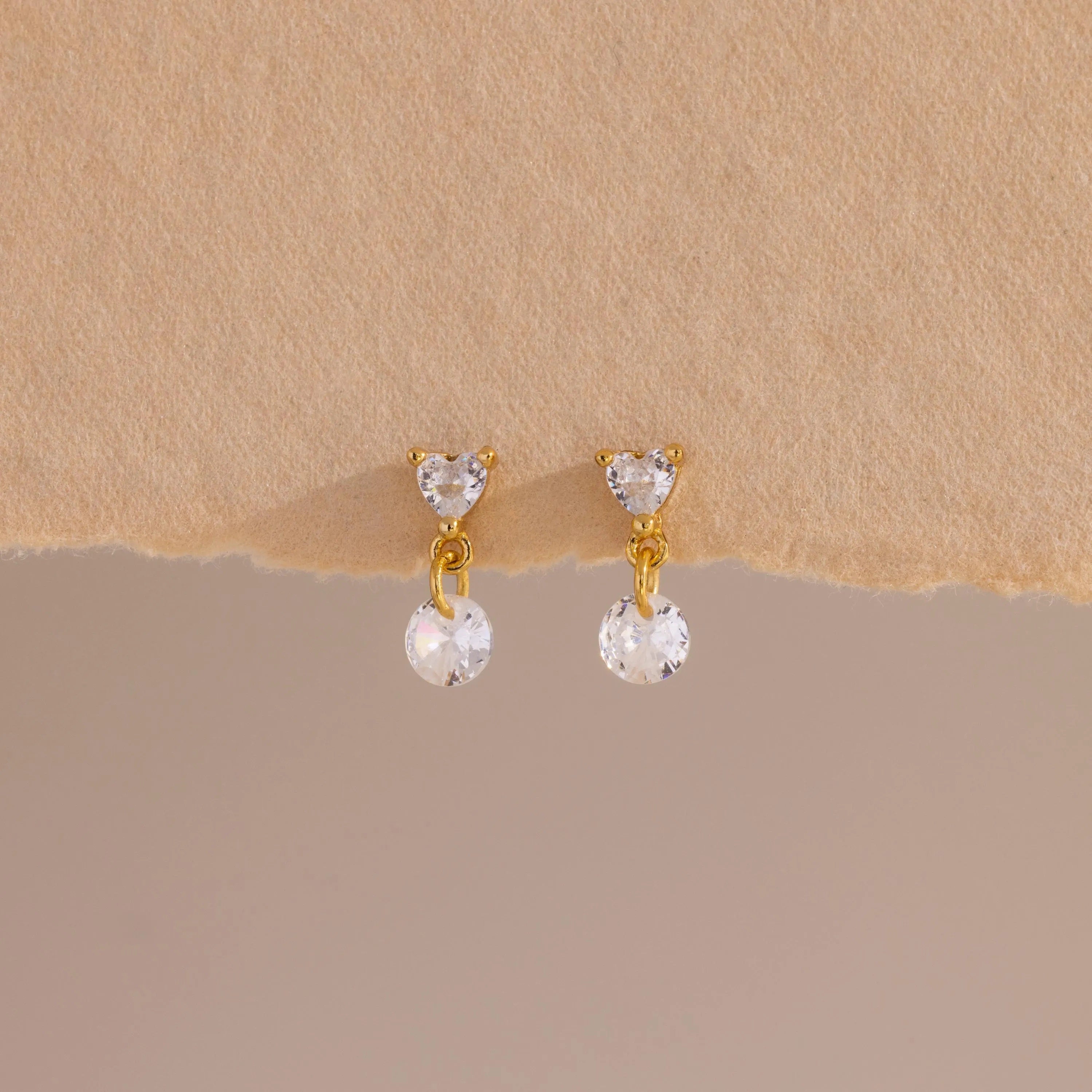 A close-up of a pair of 18k gold flat back earrings featuring heart-shaped studs with a dangling round crystal, displayed against a soft beige textured surface.