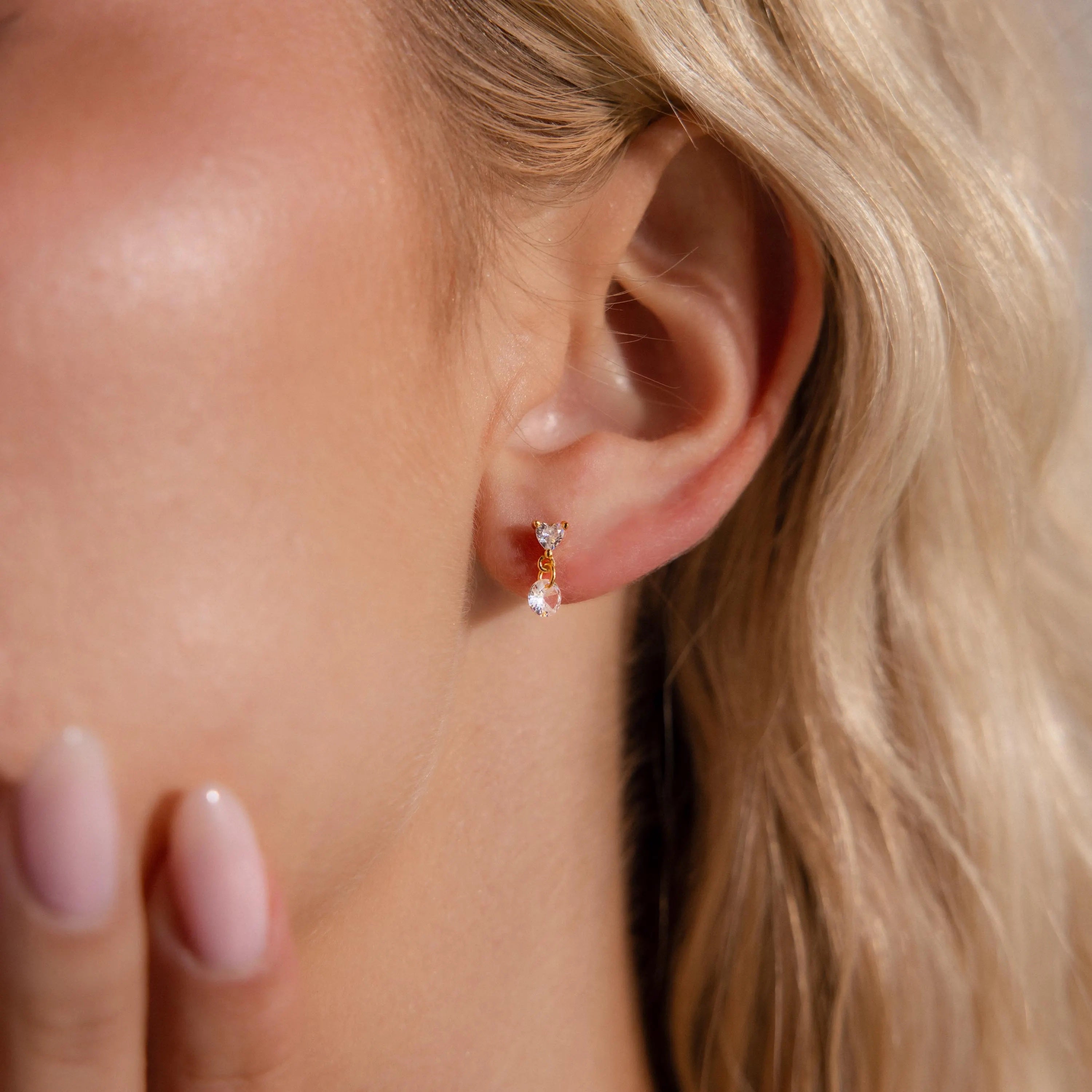 A close-up of a woman's ear showcases Heart Drop Flatback Studs, with her hand softly touching her face as the sparkling heart-shaped earrings catch the light.