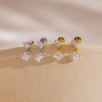 A side-by-side shot of two pairs each of the Heart Drop Flat Back Earrings in both 18k gold and sterling silver finishes, arranged on a light cream display ledge.