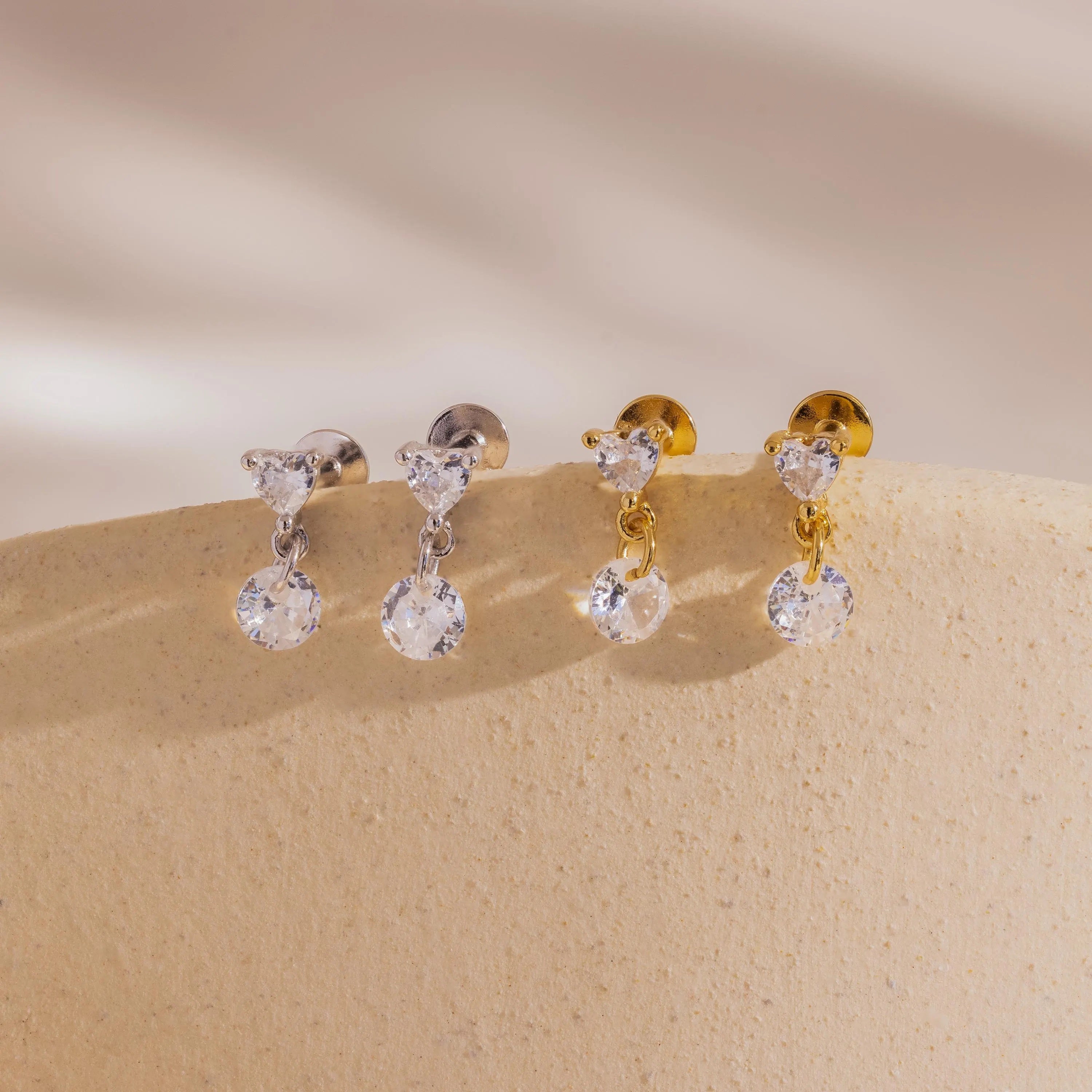 A side-by-side shot of two pairs each of the Heart Drop Flat Back Earrings in both 18k gold and sterling silver finishes, arranged on a light cream display ledge.