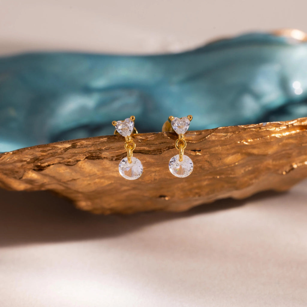 The gold Heart Drop Flat Back Earrings placed on a textured golden prop with a blurred turquoise ceramic piece in the background for an elevated aesthetic.