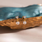 The gold Heart Drop Flat Back Earrings placed on a textured golden prop with a blurred turquoise ceramic piece in the background for an elevated aesthetic.