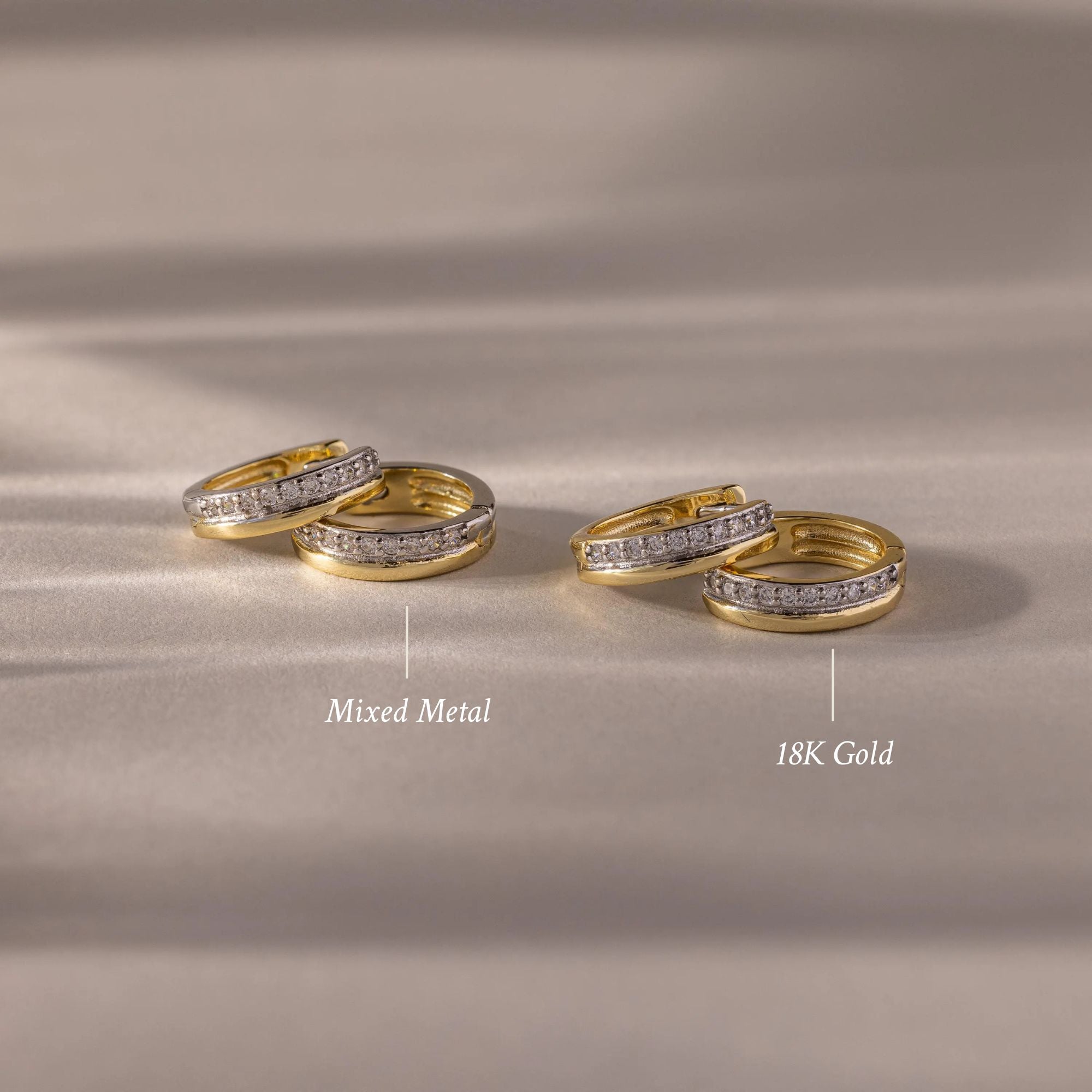 Two pairs of gold and silver Half Pave Huggies, adorned with sparkling CZ diamonds, are elegantly displayed on a soft beige surface.